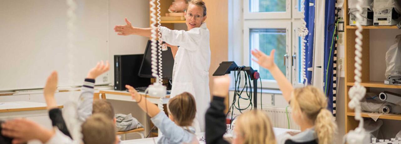 En forskare på ett universitet står och förklarar något genom att räcka ut armen och elever från årskurs två lyssnar och räcker också ut armen.