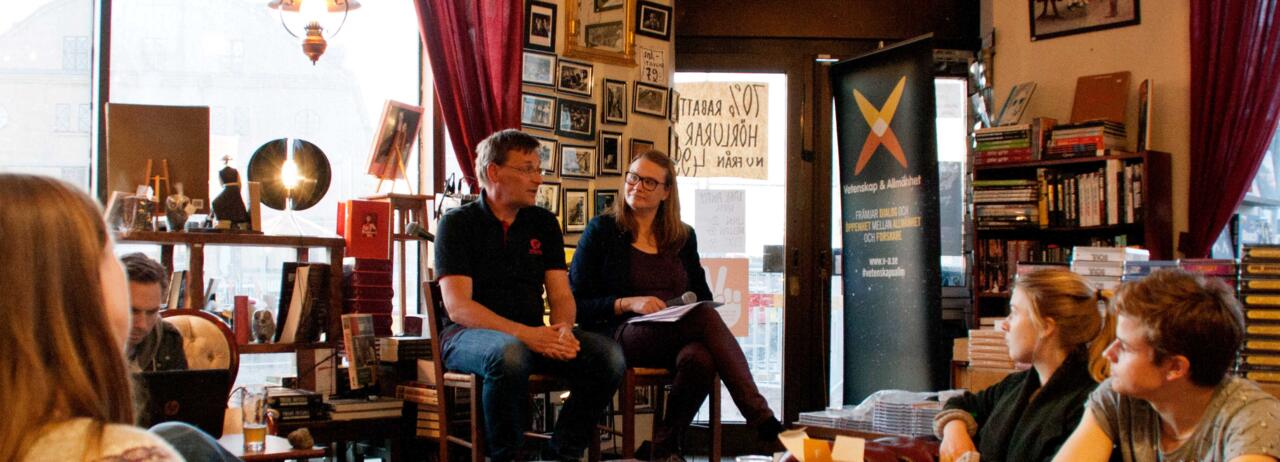 En forskare och en moderator på två stolar framför publik på ett cafe